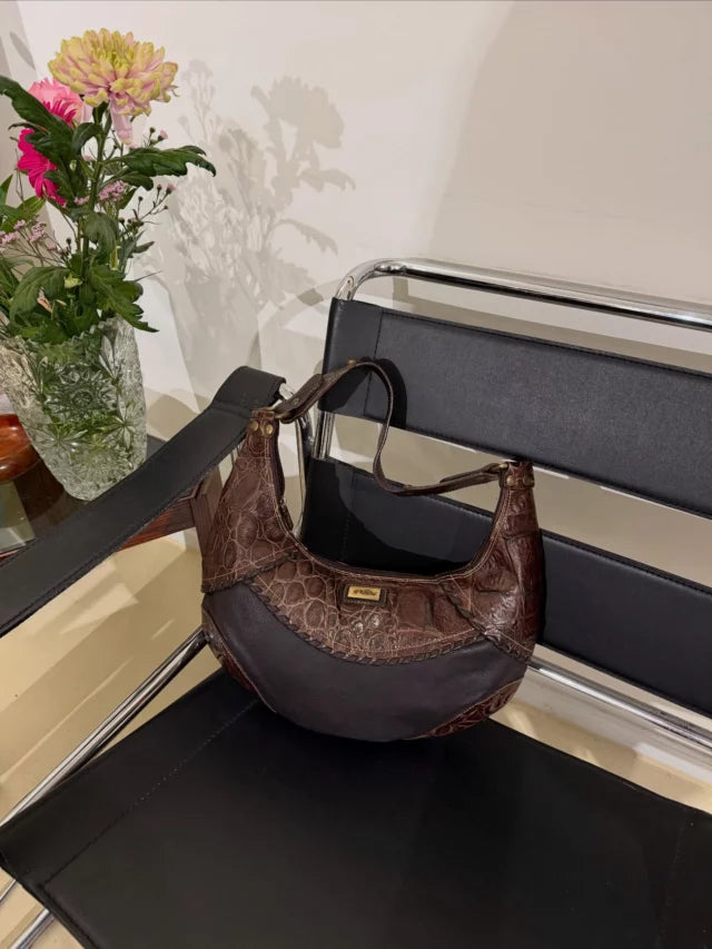 Dark brown shoulder bag displayed on a black chrome-framed chair beside flowers.